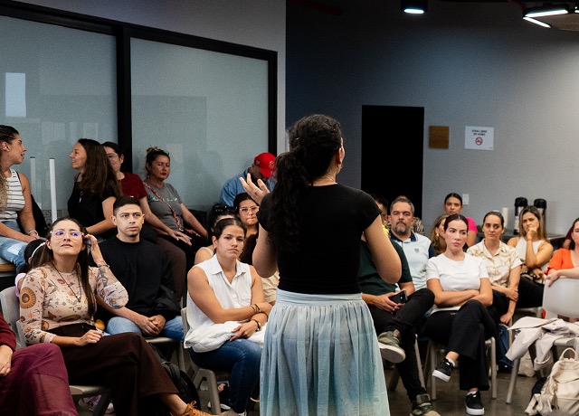 Familias sentadas en círculo escuchan a una facilitadora en un encuentro de la Escuela de Familias.
