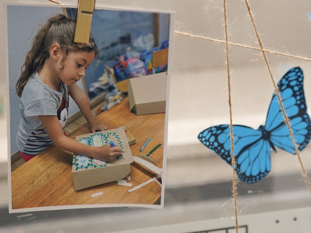 Una foto de una niña dibujando, colgada con pinzas junto a una mariposa de papel.
