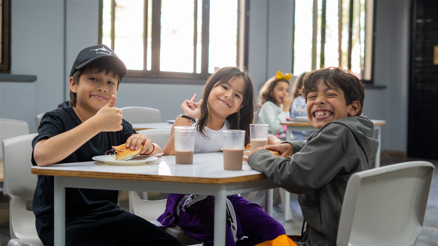 Tres estudiantes sonríen mientras comen en el restaurante escolar