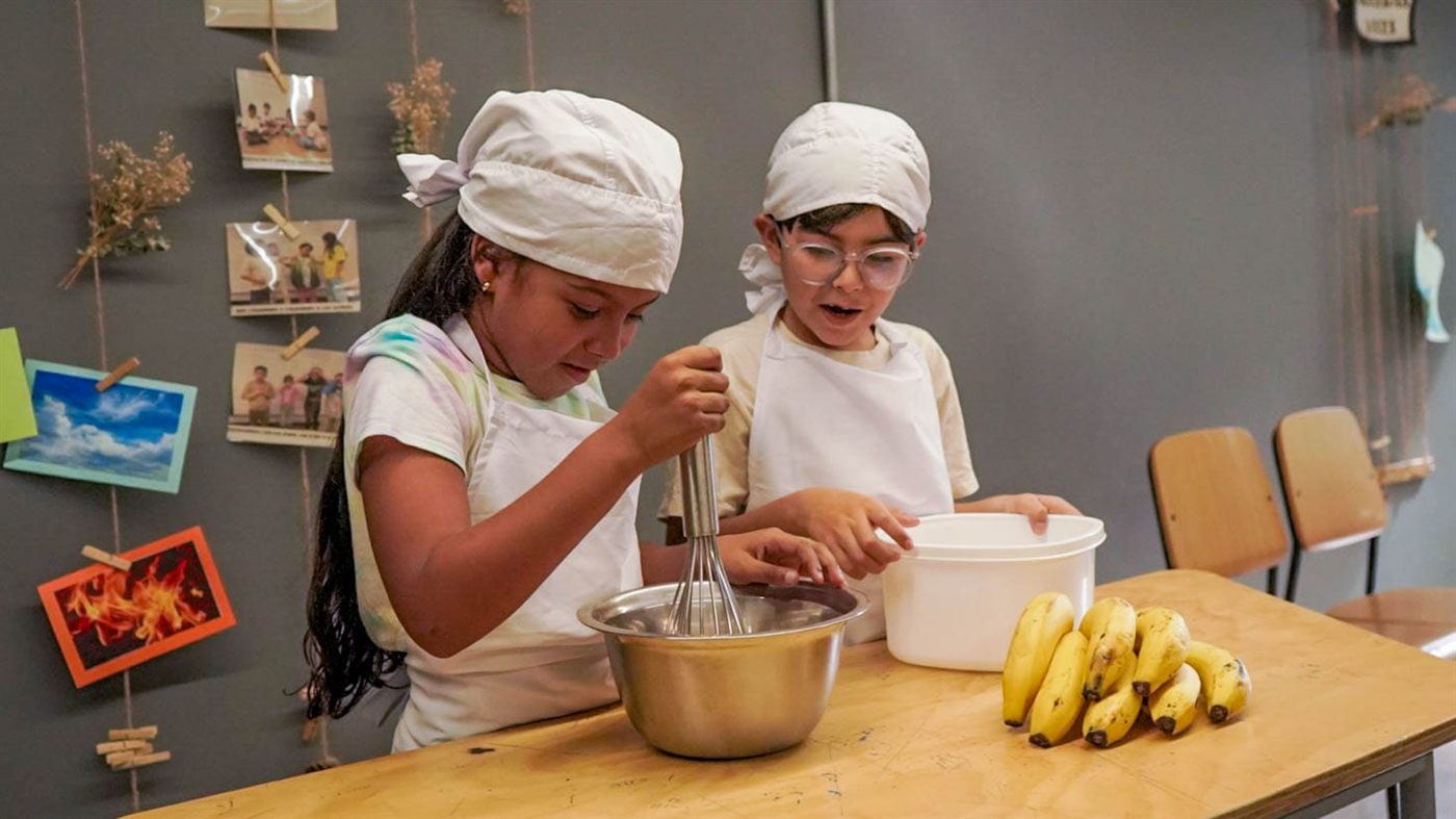 Una niña y un niño con gorro de cocina preparan una receta con bananos